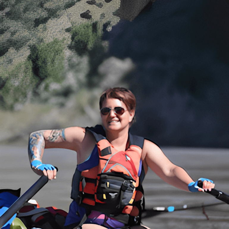 Person smiling while rowing on a raft, wearing a life jacket and sunglasses, with a rocky landscape in the background.